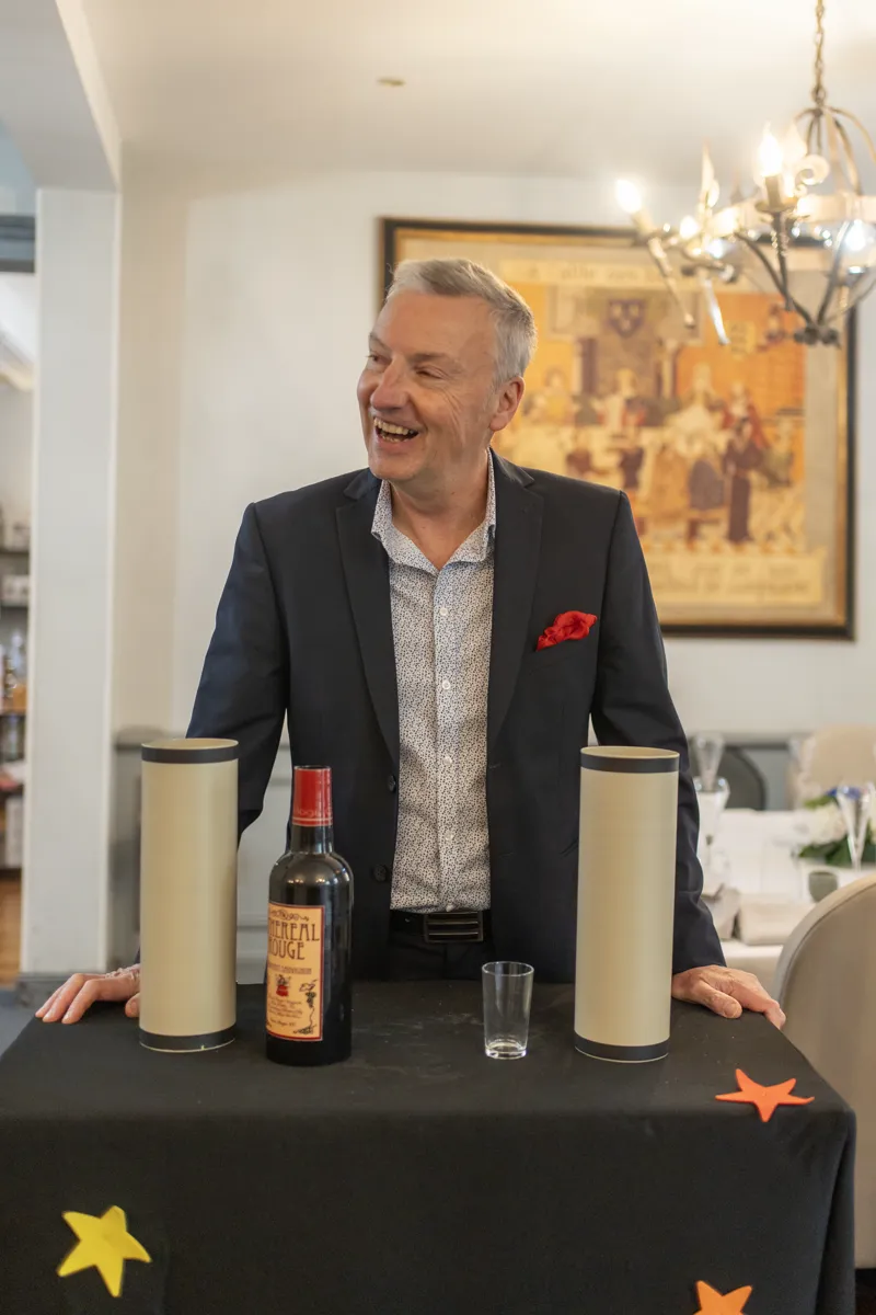 Francis Darcy derrière sa table de magie avec bouteille, cylindres et verre, photo professionnelle